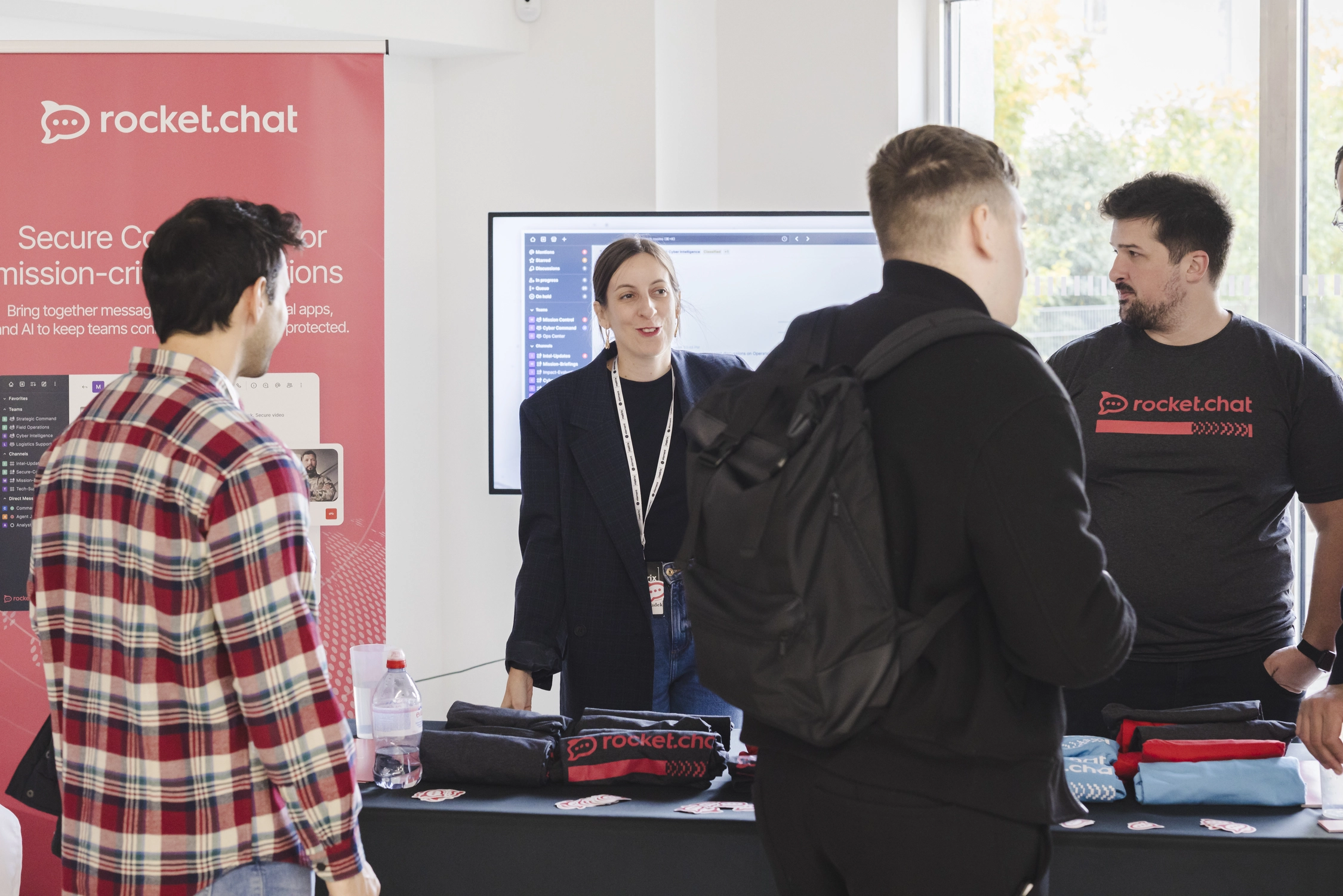 A picture of the booth of one of the Conference sponsors, Rocket.Chat, staffed by two people. Two attendees are standing in front of the booth and asking questions to the staff. On the booth table there are Rocket.Chat branded T-shirts folded.